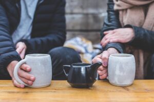 Two people sitting together at a wooden table holding ceramic mugs over tea.