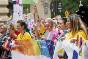 A group of smiling people marching in a parade holding trans and progress pride flags.